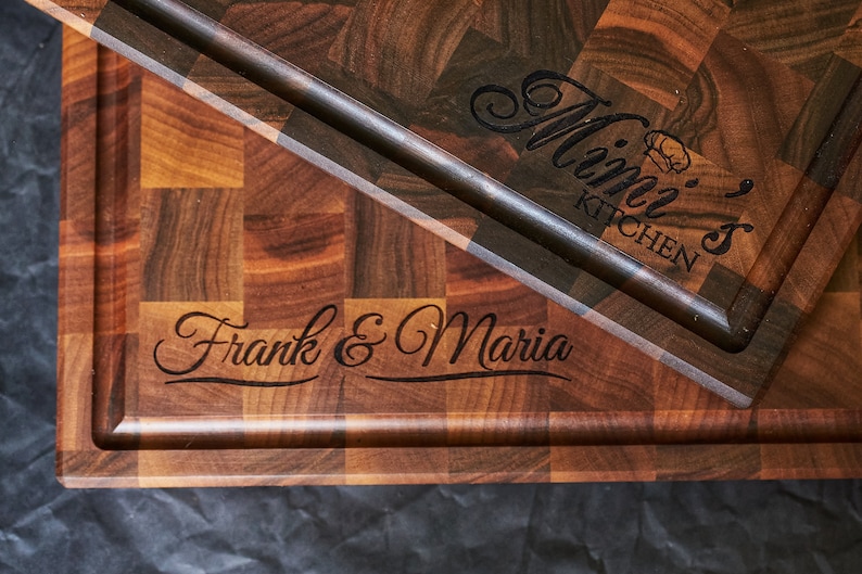 Puede incluir: Dos tablas de cortar de madera con texto grabado. La tabla superior tiene "Mimi's Kitchen" grabado. La tabla inferior tiene "Frank & Maria" grabado.