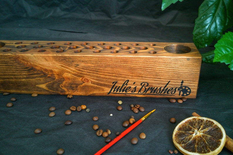 Pu&ograve; includere: Un portapennelli in legno marrone con numerosi piccoli fori e un'apertura circolare pi&ugrave; grande. Il testo "Julie's Brushes" e un logo a tavolozza sono visibili. Un pennello con manico rosso &egrave; posizionato vicino.
