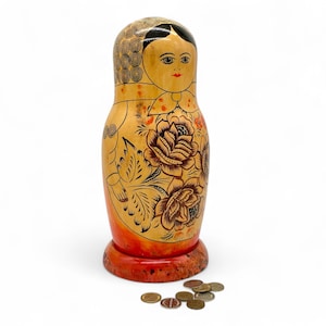 Puede incluir: Una muñeca rusa tradicional, o matrioshka, pintada con diseños florales en tonos rojos, amarillos y negros. La muñeca está hecha de madera y tiene una forma redonda. Hay varias monedas esparcidas sobre la superficie frente a la muñeca.