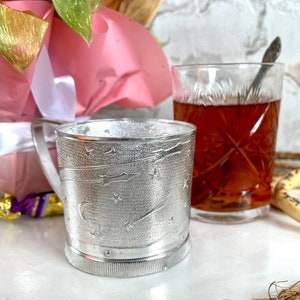 May include: A silver-colored glass cup with a handle, featuring an embossed design of stars and shooting stars. The cup is next to a clear glass with a dark liquid and a spoon.