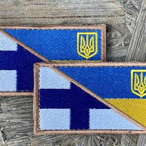 Könnte beinhalten: Zwei rechteckige, gestickte Aufnäher. Ein Aufnäher zeigt die finnische Flagge mit einem blauen Kreuz auf weißem Grund und das ukrainische Wappen. Der andere Aufnäher zeigt die finnische und die ukrainische Flagge.
