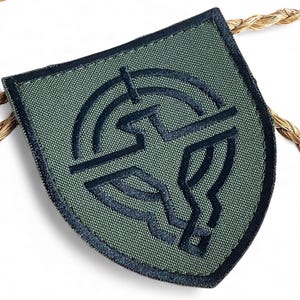 Peut inclure: Écusson brodé vert et bleu marine en forme de bouclier. L'écusson présente un emblème bleu marine au centre, avec un motif circulaire et des formes géométriques. L'écusson est probablement destiné aux vêtements ou aux accessoires.