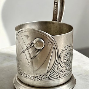 Puede incluir: Taza plateada con asa y un diseño decorativo que presenta un satélite y estrellas. La taza tiene una base con un diseño floral.