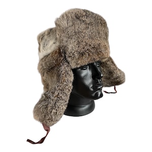 May include: A brown and tan fur hat with ear flaps and a top. The hat is displayed on a black mannequin head. The hat has a soft, textured appearance and is secured with brown ties.