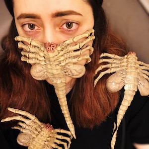 Puede incluir: Una persona que lleva una camisa negra con una máscara facial y dos piezas decorativas con forma de criaturas extraterrestres. La máscara y las piezas están hechas de un material beige y tienen una superficie texturizada detallada.