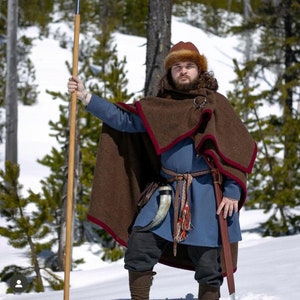 May include: A person in a historical costume stands in a snowy forest, holding a spear. They wear a brown cloak with red trim, a blue tunic, and a fur hat. A sword and drinking horn are attached to their belt. The outfit evokes a Viking or medieval theme.