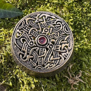May include: A round, antique-style bronze-colored metal box with intricate Celtic knotwork and a central red gemstone. The box is set against a backdrop of vibrant green moss and foliage, creating a natural, earthy aesthetic.