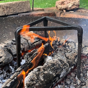 May include: A close-up shot of a fire pit with burning logs and a metal grate. Flames are visible, with a backdrop of green foliage and brickwork. The fire pit is round and made of metal, with a black metal grate over the burning wood.