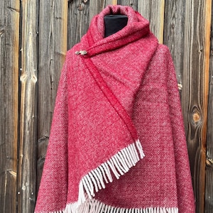 May include: A red and white woven shawl with a geometric pattern and white fringe. The shawl is draped over a mannequin, showcasing its design and texture. A decorative pin secures the shawl at the neck, adding a touch of elegance.