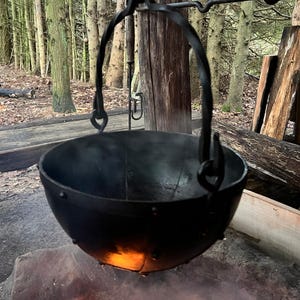 Op de afbeelding: Een zwarte gietijzeren ketel die boven een vuurplaats hangt. De ketel hangt aan een smeedijzeren arm, met een bos op de achtergrond. Vlammen en rook stijgen op van het vuur beneden.