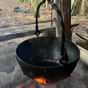 May include: A black, hand-forged iron cauldron suspended over a fire. The cauldron has a rounded bowl shape with rivets and a curved handle. The fire below casts an orange glow on the metal. The background shows a forest setting.