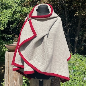 Puede incluir: Una capa gris con estampado de espiga con ribete y capucha rojos. La capa se abrocha con un broche decorativo. La capa se exhibe sobre una cabeza de maniquí y un soporte negros, sobre un fondo de follaje verde.