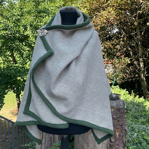May include: A gray and white herringbone patterned cloak with a green trim and hood. The cloak is fastened with a silver-colored brooch. The cloak is displayed on a black mannequin head, outdoors, with a natural green background.