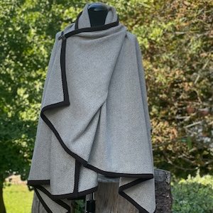 May include: A gray herringbone patterned cape with dark brown trim. The cape is draped over a mannequin, showcasing its wrap-around design. The cape has a clasp closure and is made of a soft, woven material. The background is a blurred view of green foliage.