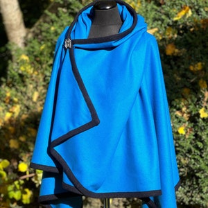 May include: A vibrant blue cape with a hood and black trim, displayed on a mannequin. The cape features a wrap-around design and is secured with a silver clasp. The background is a blurred view of green foliage.