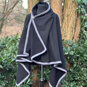 May include: A black cape with a gray trim and a decorative clasp. The cape is displayed on a mannequin outdoors, with a backdrop of trees and greenery. The cape's design features a draped neckline and a flowing silhouette.