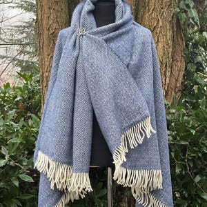 May include: A blue shawl with a diamond pattern and cream-colored fringe. The shawl is secured with a decorative silver brooch. The shawl is displayed on a black mannequin against a backdrop of trees and greenery.