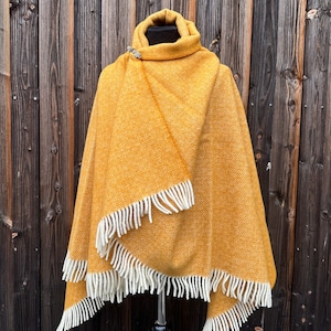 May include: Mustard yellow poncho with a geometric pattern and a high collar. The poncho has a white fringe along the edges and is secured with a silver brooch. The garment is displayed on a black mannequin.