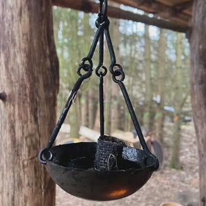 May include: A hanging black metal oil lamp with a bowl-shaped base and a wick. The lamp is suspended by a chain and three supporting arms. The background features a blurred forest scene.