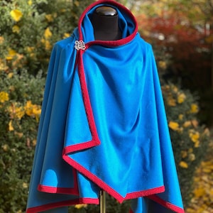 May include: A blue hooded cloak with red trim and a decorative silver clasp. The cloak is displayed on a mannequin and is made of a soft fabric. The background features fall foliage.