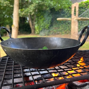 May include: A black, round wok-style cooking pan with two twisted handles, resting on a metal grill over a wood fire. Flames are visible beneath the grill. The background shows trees and a wooden post.