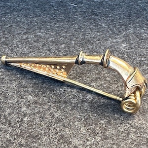 May include: A gold-toned brooch with a pin closure. The brooch features a triangular element with small perforations, a curved section, and a decorative loop. The metal has a slightly aged appearance.