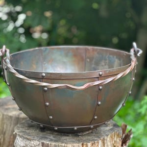 Könnte beinhalten: Eine große, runde Metallschüssel mit einer dunklen, verwitterten Patina, die sichtbare vertikale Nähte und Metallnieten an den Seiten aufweist. Die Schüssel verfügt über einen dicken, gedrehten Metallgriff, der an jeder Seite befestigt ist.