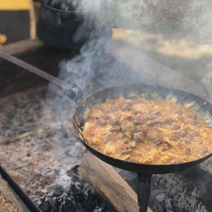May include: A cast iron skillet filled with a simmering stew over an open fire. The stew is a mix of yellow and brown ingredients, with steam rising from the pan. The skillet is supported by a metal stand.