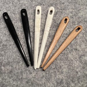 May include: A collection of six elongated hair sticks. Two are black, two are white, and two are light brown. The white sticks have carved designs. Each stick has a hole at the top.