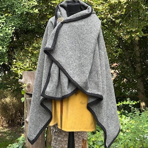 May include: A gray hooded cloak with black trim and a decorative clasp. The cloak is draped over a yellow garment on a black mannequin. The background features lush green foliage and a wooden stump.