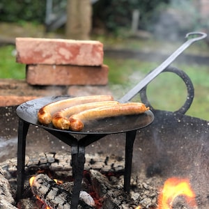 May include: Three cooked sausages on a black metal grill over a wood fire. The grill is supported by a black metal stand. Red bricks are stacked in the background. Smoke rises from the fire, and the flames are visible.