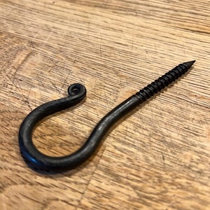 May include: A black, hand-forged iron hook with a screw end. The hook has a curved design with a looped end. The screw portion is approximately 3.8 cm long. The hook is designed for hanging items.