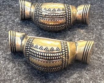 May include: Two antique brass-colored beads with intricate geometric patterns. Each bead has a barrel shape with detailed designs and vertical lines at the ends. The beads are likely for jewelry making or crafts.