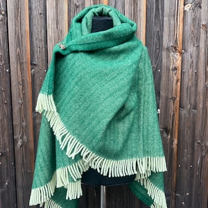 May include: A green herringbone-patterned shawl with a cream-colored fringe. The shawl is draped over a black mannequin and secured with a decorative pin. The background is a wooden wall.