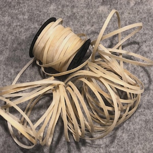 May include: A spool of natural-colored raffia ribbon with a black core. The ribbon is unwound, with some strands scattered around the spool. Ideal for crafting, gift wrapping, and decorative purposes.