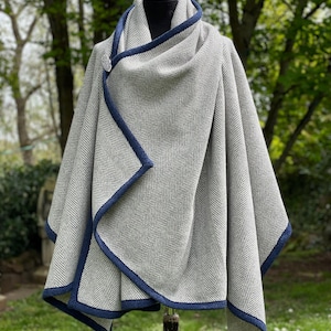 May include: A gray herringbone pattern wrap with a navy blue trim and a decorative button closure. The wrap is draped over a mannequin, showcasing its design and texture. The background features lush greenery.