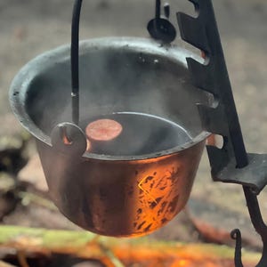 Puede incluir: Un caldero metálico rústico suspendido sobre una fogata. El caldero contiene un líquido oscuro con una rodaja de comida flotando. La escena evoca la cocina al aire libre y el camping.