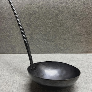 May include: A hand-forged, black metal ladle with a twisted handle. The bowl is round and deep, perfect for serving soups or stews. The ladle is made of iron and has a rustic, handcrafted appearance.