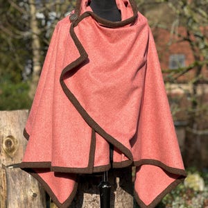 May include: A coral-colored hooded cape with dark brown trim and a single button closure. The cape is displayed on a black mannequin and appears to be made of a soft, wool-like material. The hood and edges are accented with the dark brown trim.