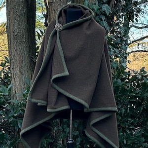 Może przedstawiać: Brązowa peleryna z kapturem, zielonym wykończeniem i ozdobnym zapięciem. Peleryna jest prezentowana na manekinie, podkreślając jej drapowany design. Tkanina wydaje się być grubą wełną, odpowiednią na chłodniejsze dni.