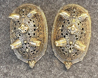 May include: Two ornate, gold-colored metal brooches with intricate, openwork designs. Each brooch has an oval shape with a central cross motif and four raised, rectangular accents. The brooches are likely vintage or antique, and are displayed on a gray surface.