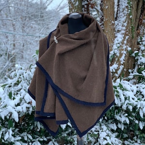 May include: A brown wool cape with a dark blue trim and a decorative silver brooch. The cape is draped over a black mannequin head. The background features a snowy outdoor scene with trees and bushes.
