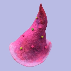 Puede incluir: Un sombrero de fieltro rosa hecho a mano con una parte superior puntiaguda y un ala ancha y con volantes. El sombrero está adornado con pequeñas bolas de fieltro de colores en tonos rosa, verde y amarillo. El sombrero está sobre un fondo lila claro.