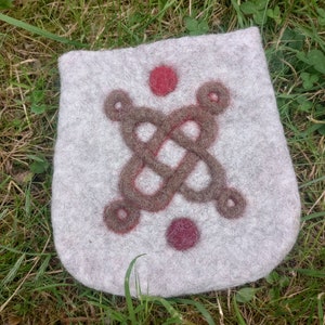 May include: A gray felt pouch with a brown Celtic knot design and two red circles. The pouch is lying on a bed of green grass. The pouch is a soft, handmade item.