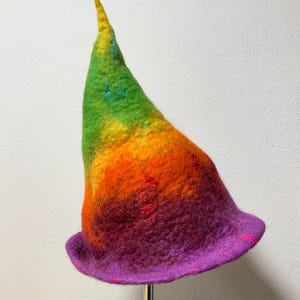 Może przedstawiać: Ręcznie robiony kapelusz filcowy w tęczowych kolorach, od zielonego do fioletowego. Kapelusz ma wysoką, spiczastą koronę i szerokie rondo. Kapelusz jest prezentowany na metalowym stojaku, ma około 30 cm wysokości.