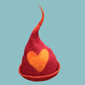 Puede incluir: Un sombrero de fieltro hecho a mano en tonos rojos y naranjas. El sombrero presenta un gran corazón naranja en la parte delantera y una punta alta y curvada. El borde y la punta del sombrero son de un naranja más brillante. El sombrero está sobre un fondo azul claro.