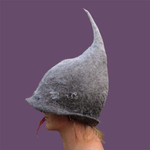 Può includere: Un cappello di feltro grigio con una corona alta e appuntita e un'ampia tesa. Il cappello ha un design stravagante da favola, con piccole decorazioni floreali. Una foglia rossa è posta vicino alla bocca di chi lo indossa. Il cappello è su sfondo viola.