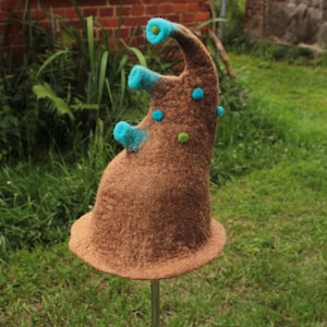 Può includere: Un cappello stravagante in feltro marrone dalla forma organica unica. Il cappello presenta tre protuberanze turchesi ed è adornato da pois in feltro turchese e verde. Il cappello è esposto all'aperto, sullo sfondo di erba verde e un muro di mattoni.