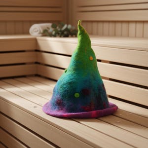 Puede incluir: Un gorro de sauna colorido con forma cónica. El gorro está hecho de fieltro y presenta un degradado de colores, incluyendo verde, azul y morado. Tiene un ala ancha y está decorado con pequeños acentos redondos. El gorro está sobre un banco de madera.