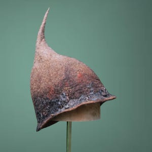 Könnte beinhalten: Ein brauner Filzhut mit einer langen, gebogenen Spitze. Der Hut hat einen dunklen Rand und wird auf einem Ständer vor grünem Hintergrund präsentiert. Der Filz hat ein strukturiertes Aussehen mit Farbvariationen.